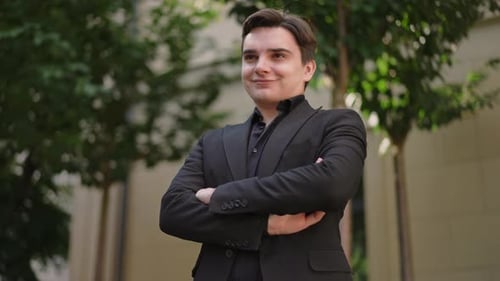 A Young Businessman in a Dark Suit Grinning and Crossing His Arms in Front of Him Stands Near a