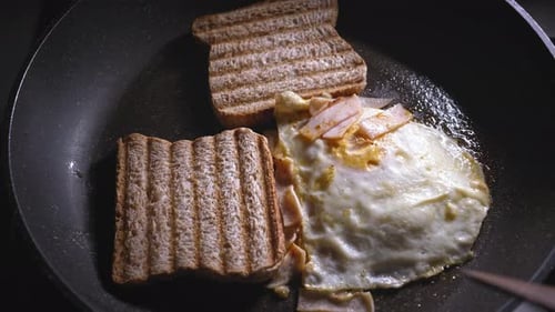 A close up shot of toasted bread being placed into a frying pan alongside a fried egg topped with ha