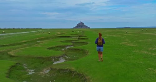 A Female with a Backpack Visits the Mont Saint Michel Castle While Walking Through Green Meadows