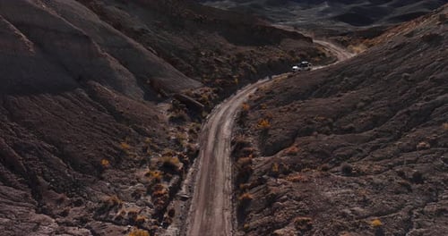 Descending aerial white off road SUV driving on dirt road through desert landscape