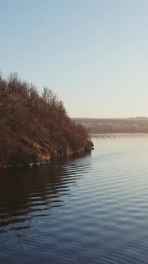 Flying over the water. Trees on the hill near the water in winter season.