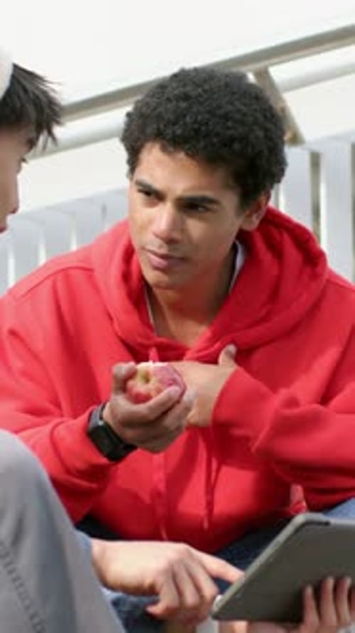 Vertical video: Tapping tablet left peer guiding red-hoodie man with bitten fruit biting on bench