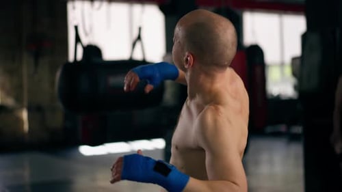Nahaufnahme in einem Boxclub Glatzköpfiger junger Boxer mit Tattoos in blauen Bandagen, Zügen, Schlägen, Schatten