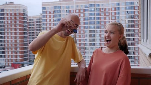 Couple Holding Keys on Balcony With Apartment Building