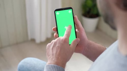 Young Man Sitting at Home Holding Smartphone Green Mockup Screen in Hand Male Person Using Chroma