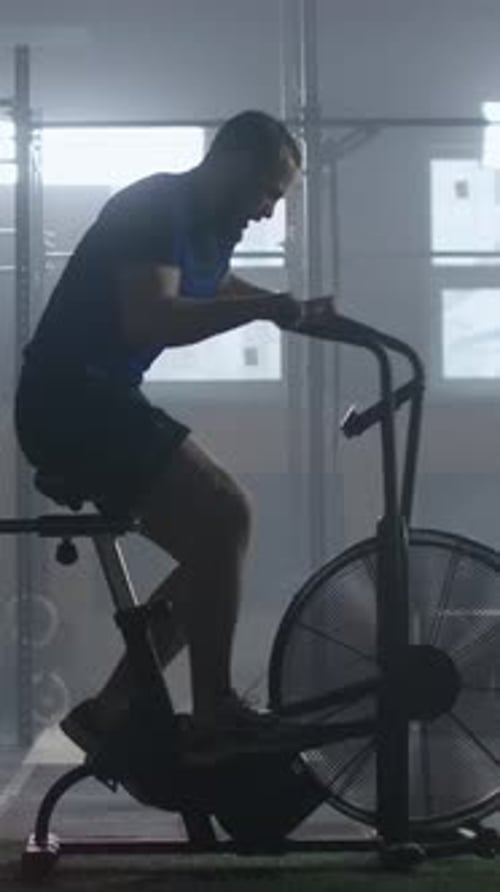 Focused Athlete Training Intensely on Stationary Air Bike in Dimly Lit Gym Pushing Endurance and