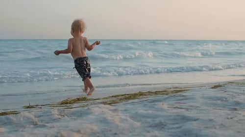 Cute Child Running To Mom At Sunset Beach. Happy Mother Playing With Baby Boy In Sea Waves At Coa...