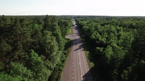 Fly over straight road surrounded by forest, truck and car approaching