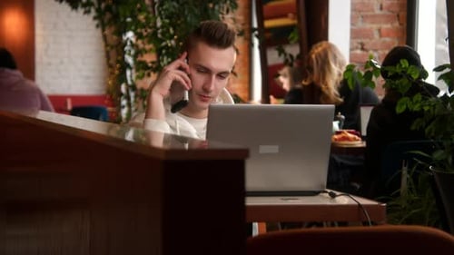 Man Talking on Phone and Using Laptop in Cafe