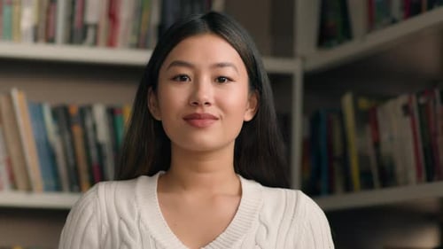 Young Asian Woman Student Posing in University Library or Bookstore with Bookshelf in Background