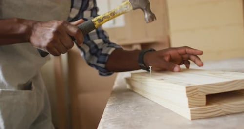 Mid section of african american male carpenter hammering nails into the wood at a carpentry shop