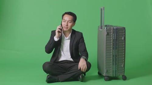 Asian Businessman Traveler With Luggage Talking On Smartphone While Sitting In The Green Screen