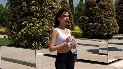 A Woman in Sportswear with Wireless Headphones Drinking Water in a Park