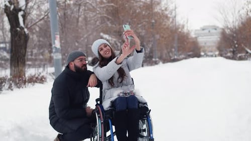 Disabled Woman in Wheelchairs Take Selfie with Man on Smartphone at Winter Park Front View Disabled