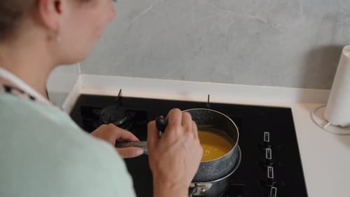 A view from above and behind the woman, she stirs food in a pot on the stove in slow motion.