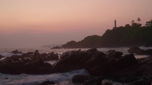 Lighthouse Stands on Green Hill at Dusk Overlooking Ocean with Waves Crashing on Rocky Shore Pink
