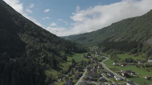 Drone shot flying in between the mountains with a small village on the ground on a sunny and cloudy
