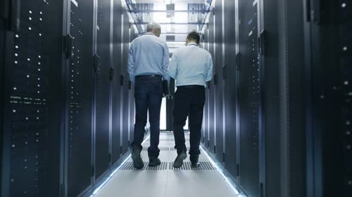 Two Server Technician Working in Data Center. One Uses Tablet Computer. They Walk Through Rows of S