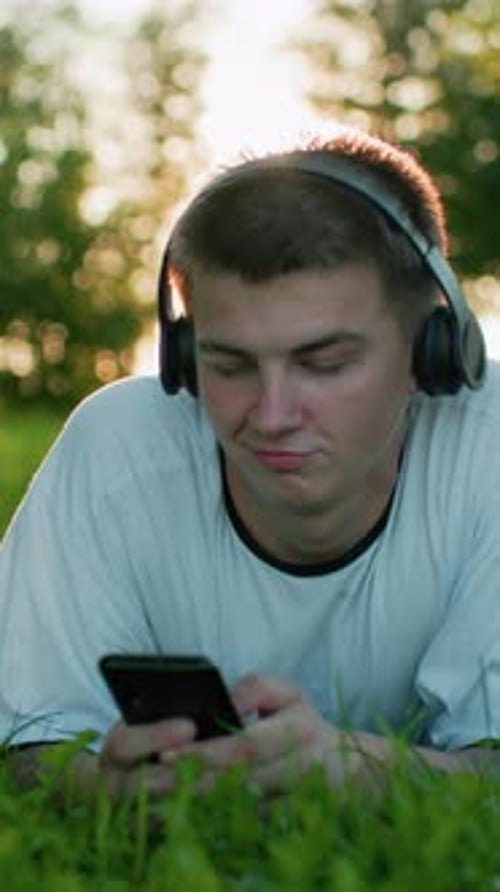 Man Lying in Grass, Wearing Headphones, Using Phone