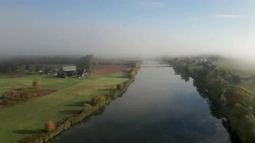 Morning fog hangs over a smooth river and peaceful green farms along the banks. Aerial view of rural