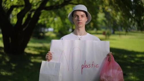 Un jeune homme coiffé d'un chapeau de soleil regarde la caméra et tient une affiche sans plastique dans ses mains pendant