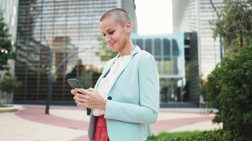 Modern business woman with short hair happily typing messages on her mobile phone outside the office