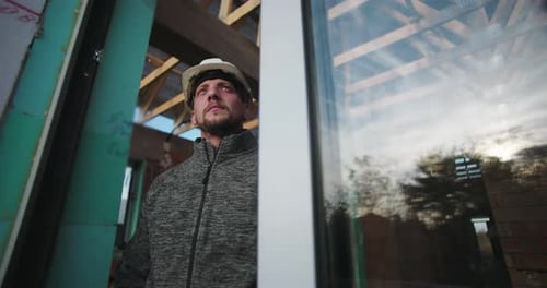 Young construction worker in hard hat, reflective expression, standing by glass and wood framework