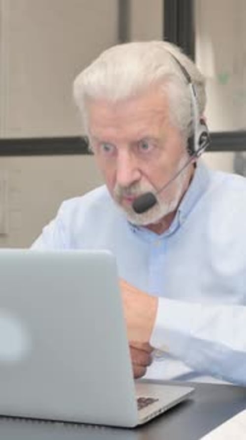 Senior Man with Headset Doing Video Chat on Laptop in Call Center
