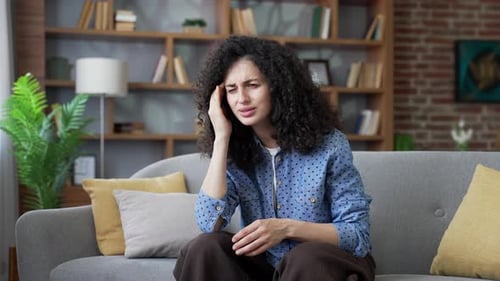 Unhealthy young woman suffering from headache sitting on sofa in living room at home. Unwell adult
