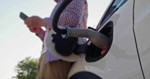 Man With Mobile Phone Stands Near Electric Car Connected to a Charging Station