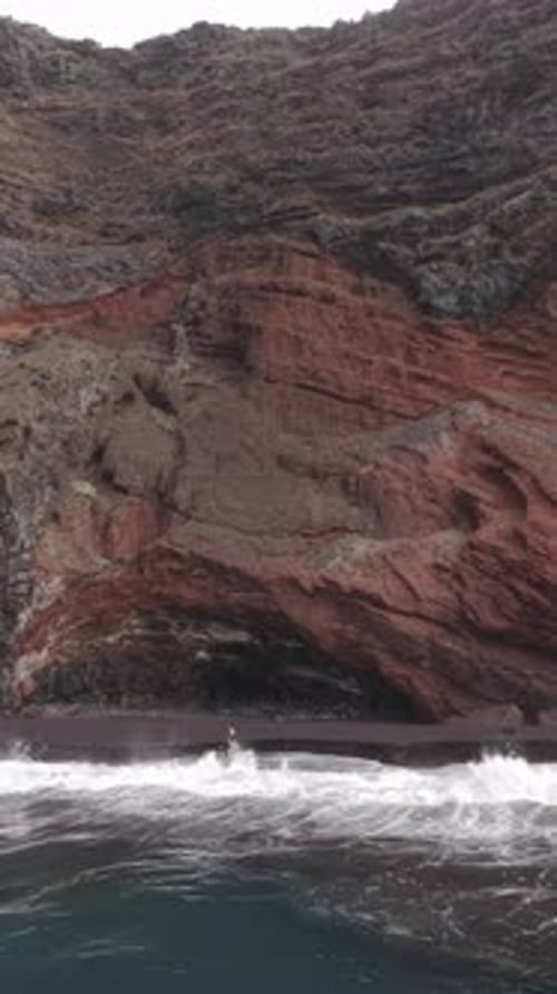 Powerful Waves Breaking Against Black Volcanic Sand in a Vertical Video Red Cliffs Towering Behind