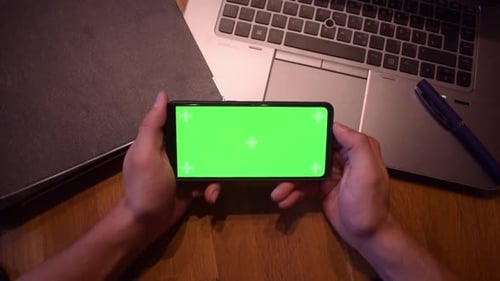 Ein
junger erwachsener Junge hält ein Smartphone mit einem Greenscreen in der Hand. Ein Video starten. Notizbuch auf dem D