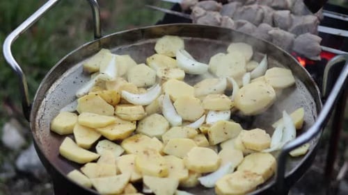 potatoes with onion are cooking in a large metal frying pan on the grill on a charcoal over an open