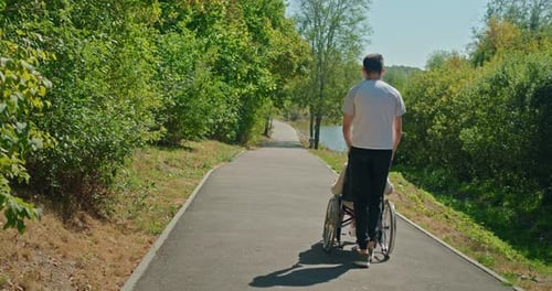 Man Pushing a Middleaged Woman in a Wheelchair Along a Forest Path on a Sunny Day Surrounded By Lush