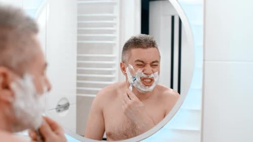 Man Shaves Face in Bathroom Mirror