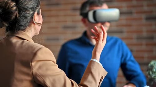 Man using a Virtual Reality headset in an office while woman explains and gesticulates