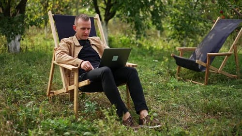 Calm relaxed freelancing man sitting in apple garden with laptop on his knees.