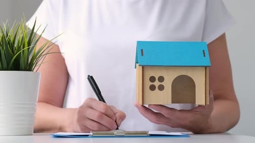 Woman Signs Paperwork, Holding a Small House Model