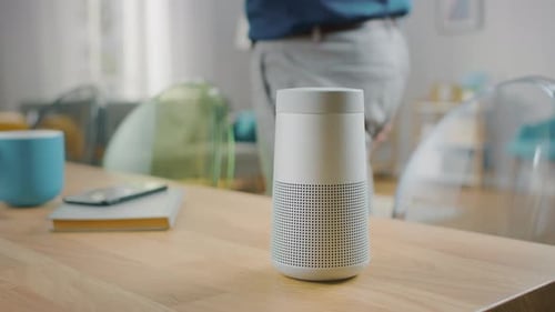 Close Up Shot of a Modern Silver Wireless Speaker Standing on a Table at Home. Man in the Backgroun