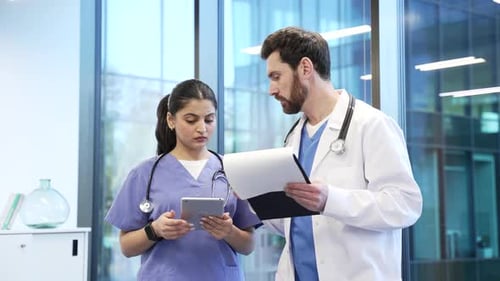 Two doctors man and woman collaborate over a tablet and folder standing in a hospital office