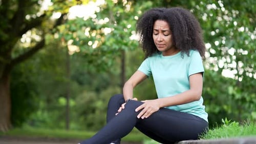 Young african american sportswoman sitting with muscle pain in city park. Black woman massaging