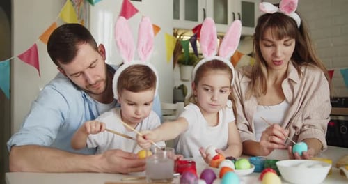 Family Wearing Bunny Ears Painting Easter Eggs Together in Bright Modern Kitchen