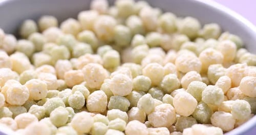 Tasty breakfast cereal falling into bowl on lilac background, closeup