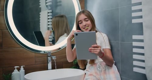 Niña bastante divertida con pelo largo en pijama cepillándose los dientes y usando una tableta por la mañana