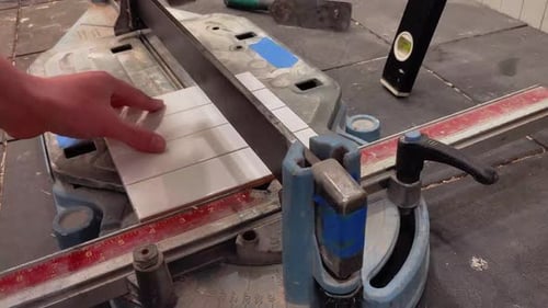 A white male construction worker uses a tile snap cutter to cut long white ceramic tile pieces for a