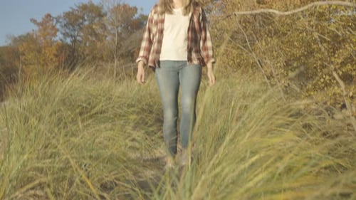 Handheld pan shot from behind dune grass to reveal a woman walking through the sand to the beach at