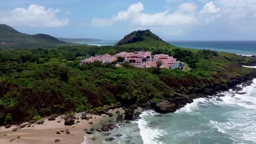 Coastal resort town with red-roofed buildings nestled by the sea, lush greenery, sunny day, aerial v