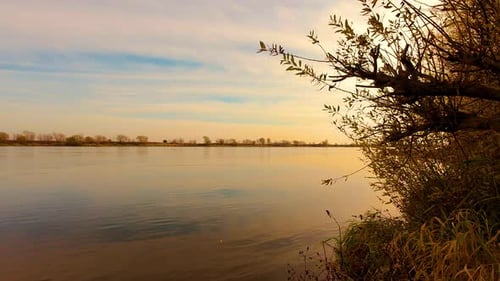 Tranquil River with Bushes Growing Along Banks Reflecting Sky with White Clouds in Rays of Sunset