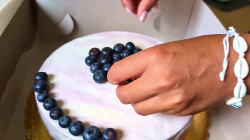 Close-up view of woman's hands with pink nails placing blueberry fruit on top of vanilla cake in car