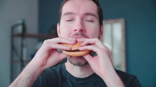 Slow motion young caucasian man bites into burger at home, cheeseburger eating enjoyment, close up p